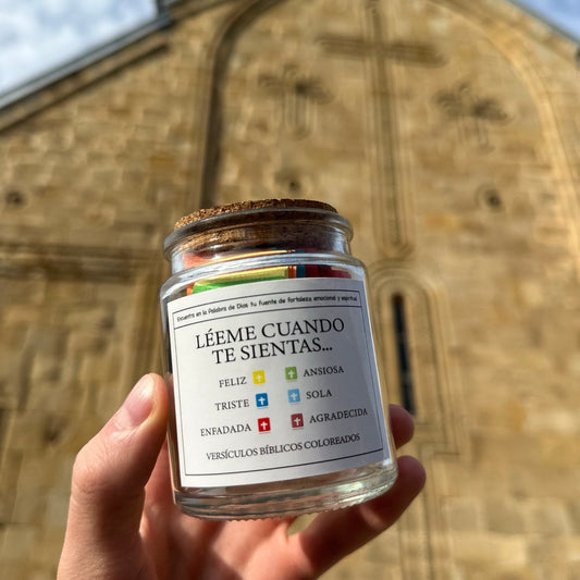 Un frasco de vidrio etiquetado "Léeme cuando te sientas..." se sostiene frente a una iglesia de piedra con una cruz. Contiene tarjetas de colores con emociones y versículos bíblicos, ofreciendo guía espiritual y apoyo emocional.