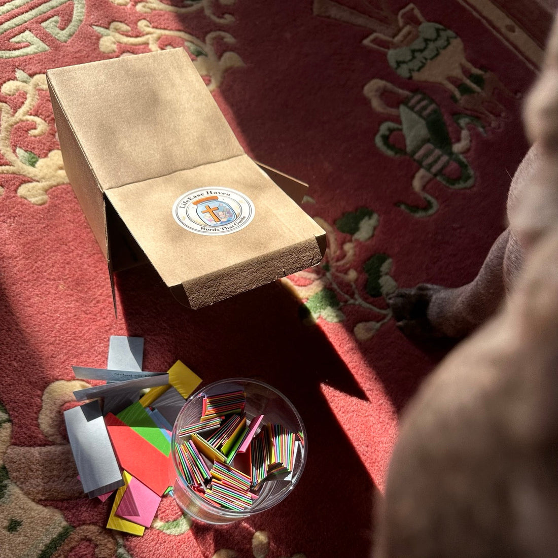 Pohled na dárkové balení LifeEase Haven: hnědá papírová krabička a skleněná Nádoba inspirace s rozptýlenými barevnými kartičkami biblických veršů na červeném koberci.